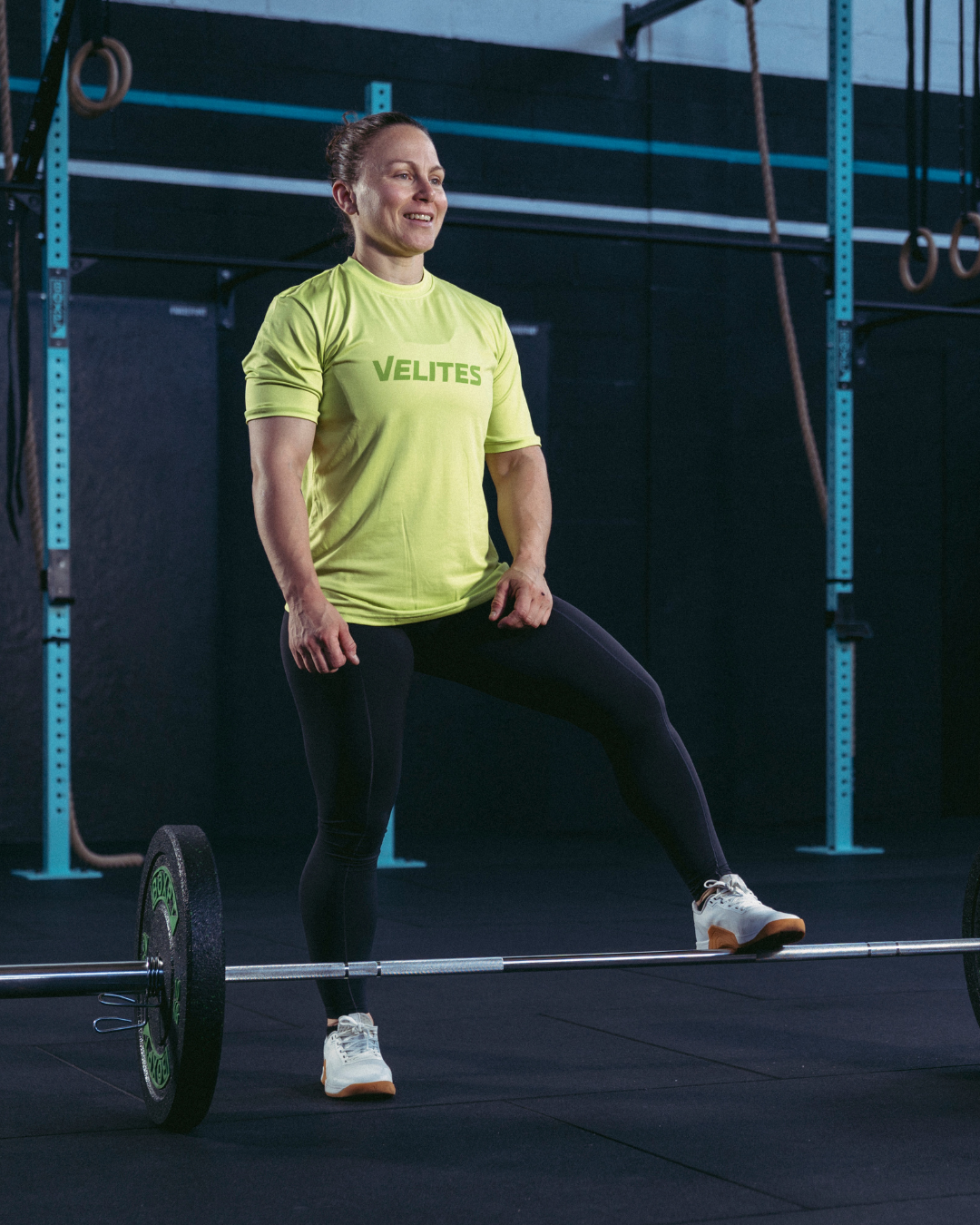Female athlete in a CrossFit box wearing a yellow Velites Holo t-shirt next to a loaded barbell