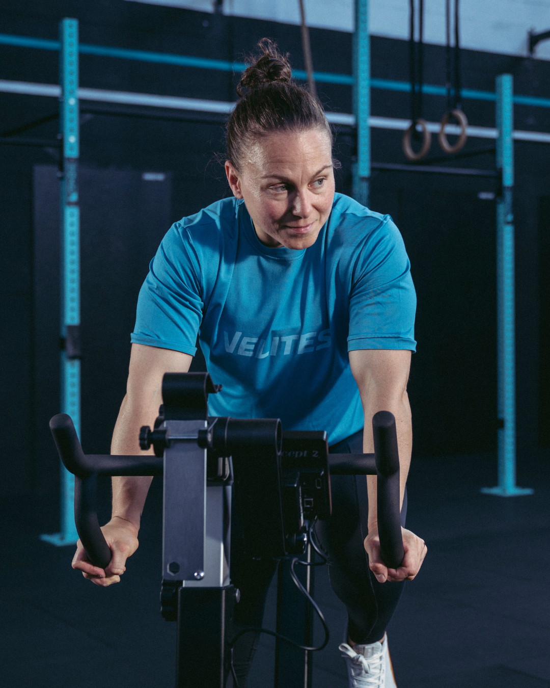 Female athlete training on a BikeErg wearing a blue Velites Holo t-shirt in a box