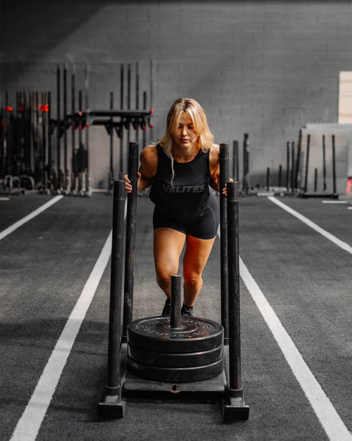 Female athlete pushing a weighted sled in a CrossFit box wearing a black Velites tank top